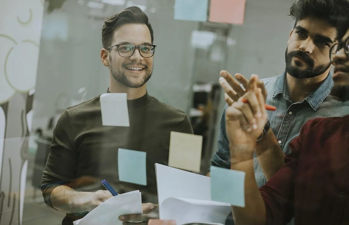 Eine Frau sitzt vor einem Laptop und bedient zugleich ein Smartphone. Auf beiden digitalen Endgeräten werden Social-Media-Plattformen angezeigt, die sie dank ihres Social-Media-Redaktionsplans strukturiert mit Content bespielt. Diverse Interaktions-Icons symbolisieren den regen Zuspruch auf ihre Posts.