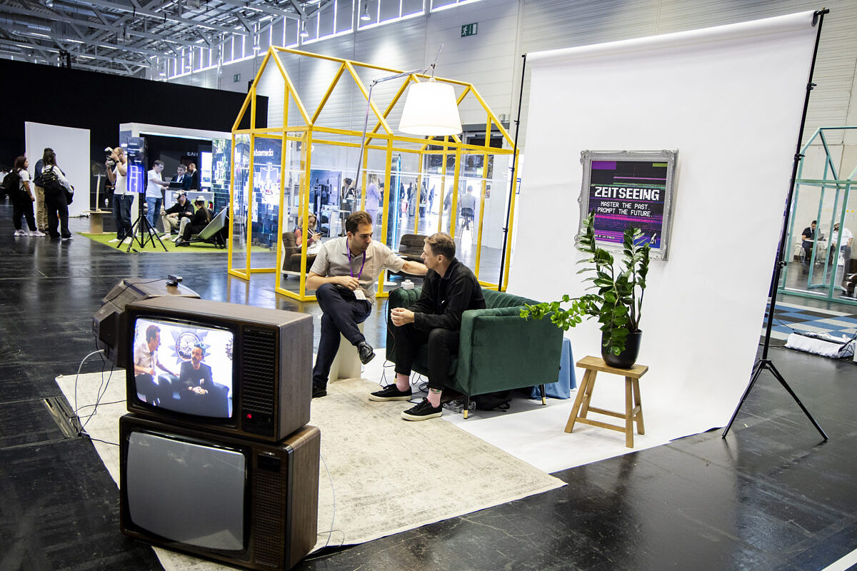 Installation „Zeitseeing“ von Jung von Matt auf der DMEXCO 2024 Zwei Männer sitzen nebeneinander in einer Halle, vor ihnen ein Röhrenfernseher.